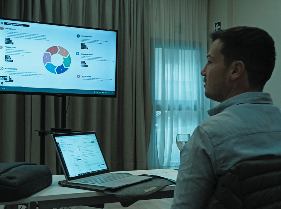 Un homme assis devant un ordinateur portable regarde un écran de présentation avec un graphique circulaire dans une salle avec rideaux.