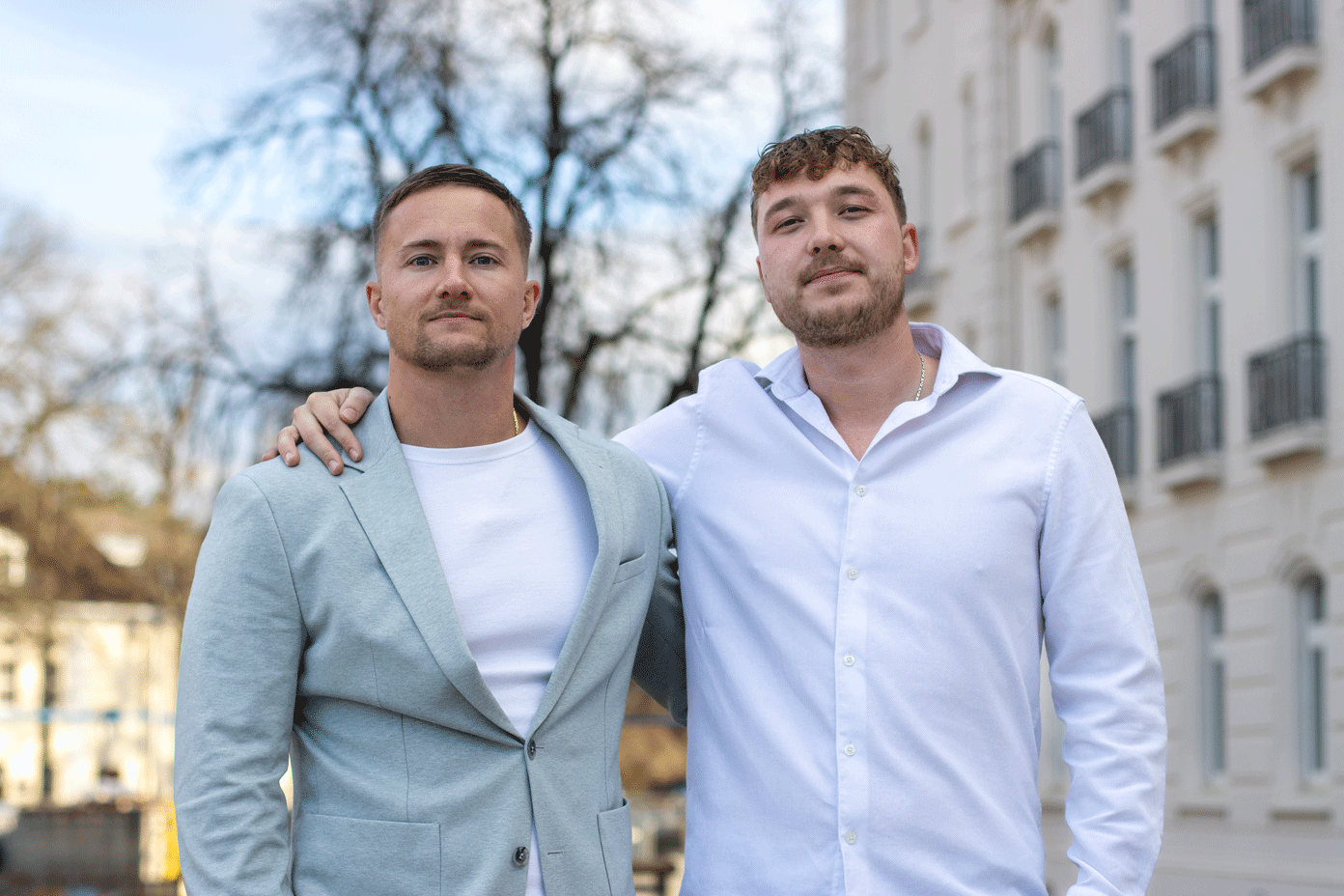 Zwei Männer stehen im Freien, einer in einem hellgrauen Blazer und einem weißen Hemd, der andere in einem weißen Hemd mit Knöpfen, und haben die Arme umeinander gelegt.