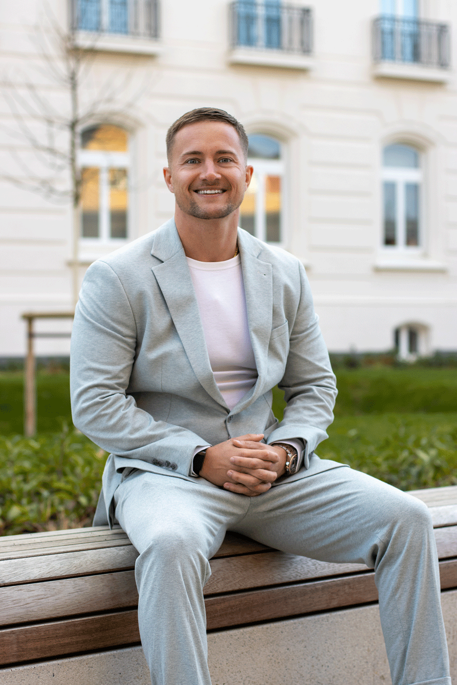 Lächelnder Mann in einem hellgrauen Anzug und weißem Hemd, der auf einer Holzbank vor einem Gebäude mit Fenstern im Freien sitzt.