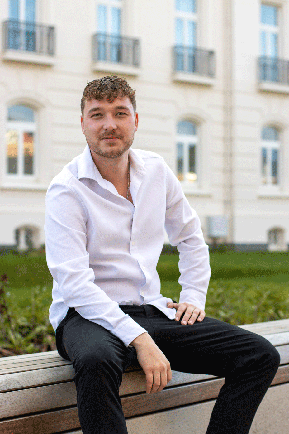 Junger Mann mit gestutztem Bart, bekleidet mit einem weißen Hemd und einer schwarzen Hose, sitzt auf einer Holzbank im Freien vor einem beigen Gebäude mit Fenstern.