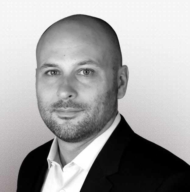 Portrait of a bald man with a beard wearing a white shirt and dark blazer against a light background.