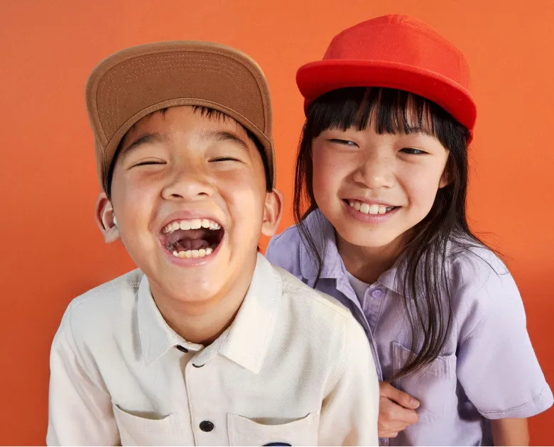 Two children smiling and wearing caps, one in a red cap and lavender shirt and the other in a brown cap and light beige shirt, against an orange background.