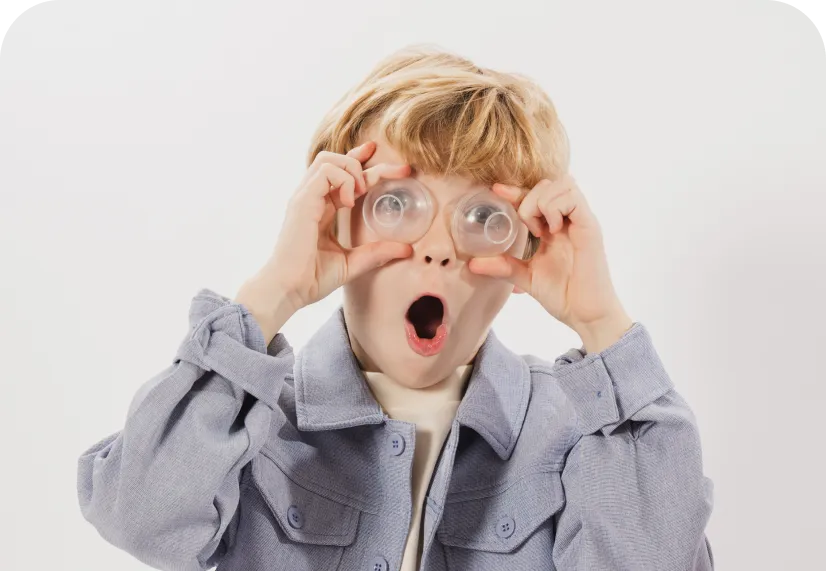 Young boy with blonde hair holding two clear suction cups over his eyes with a surprised expression.
