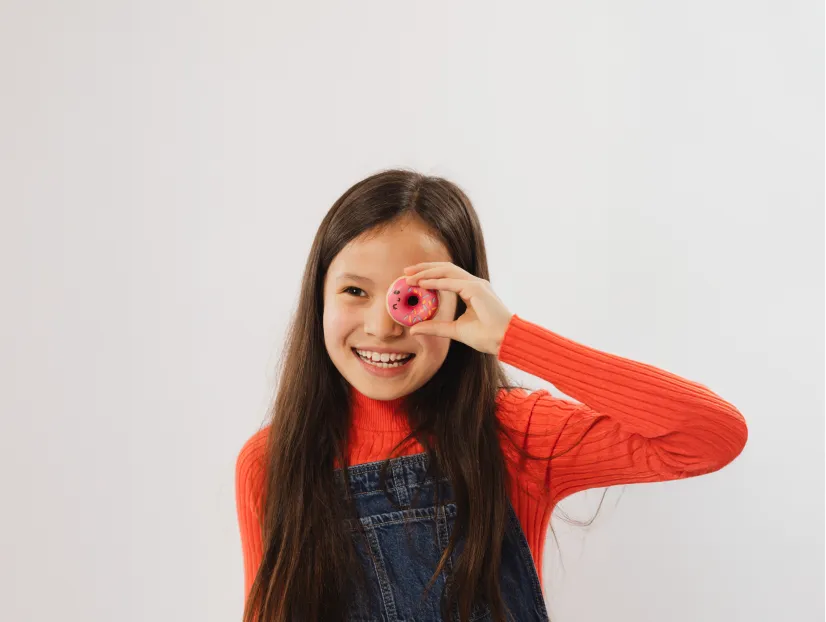 Smiling girl in a red sweater and denim overalls holding a pink donut with sprinkles over one eye.