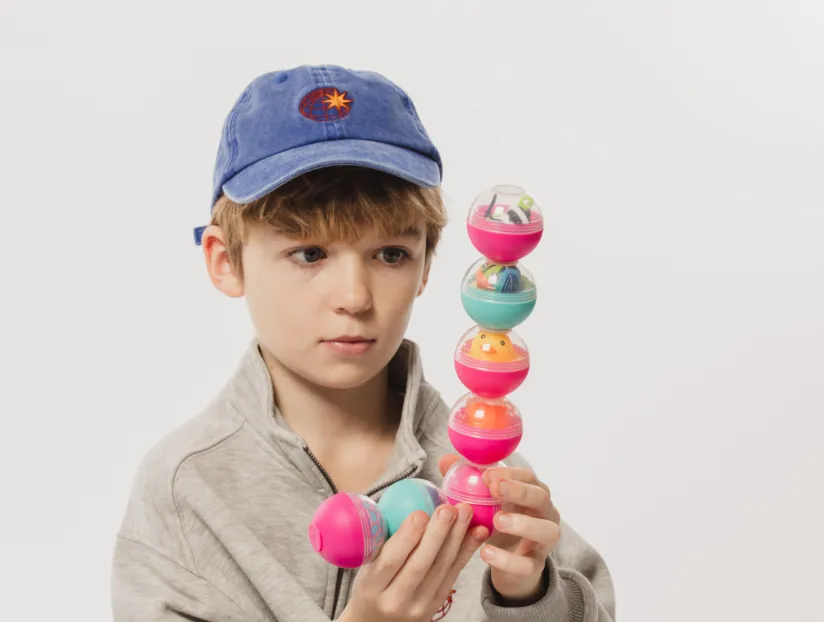 Young boy wearing a blue cap and grey jacket stacking colorful plastic capsules.