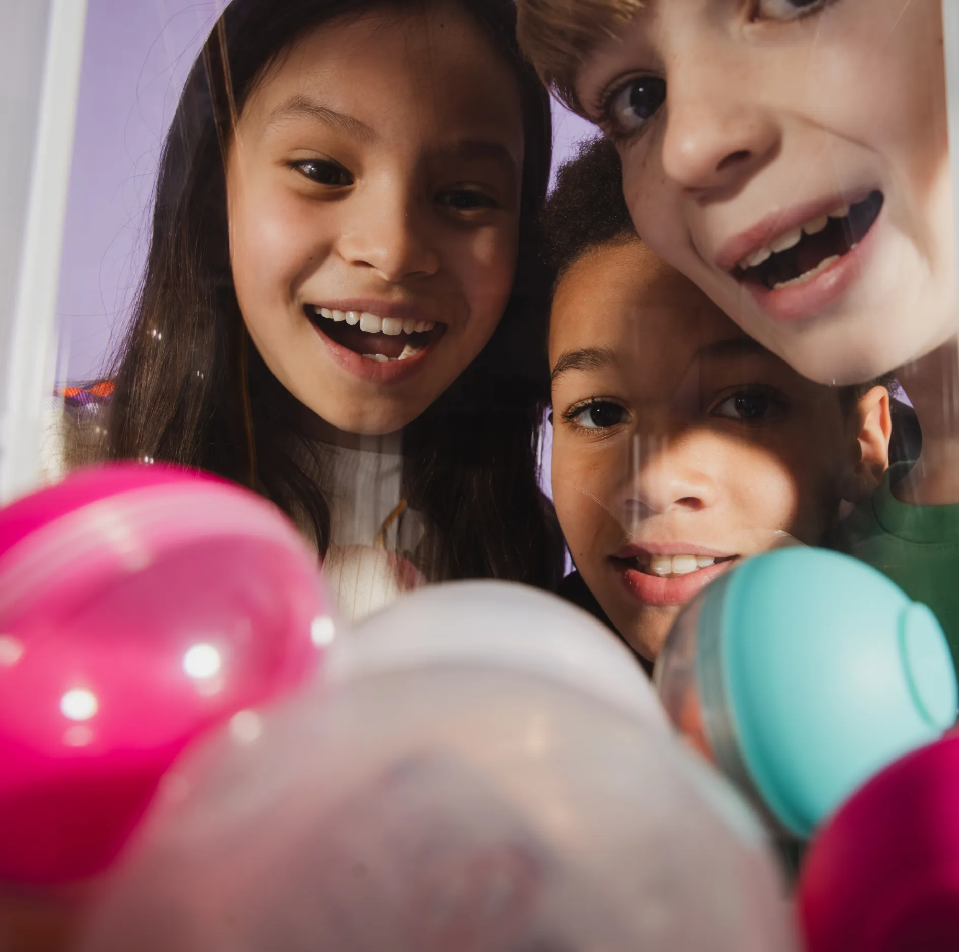 Three children looking excitedly at colorful capsule toys inside a vending machine.