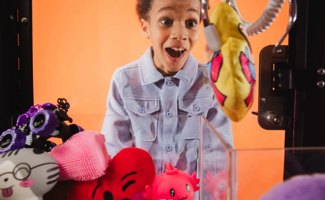 Excited young boy watching a claw machine with colorful plush toys inside against an orange background.