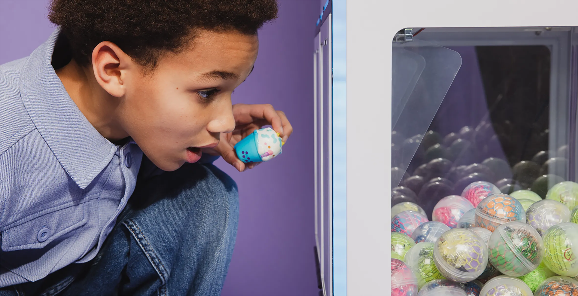 Young boy holding a small toy ice cream cone while looking excitedly into a claw machine filled with colorful capsule toys.