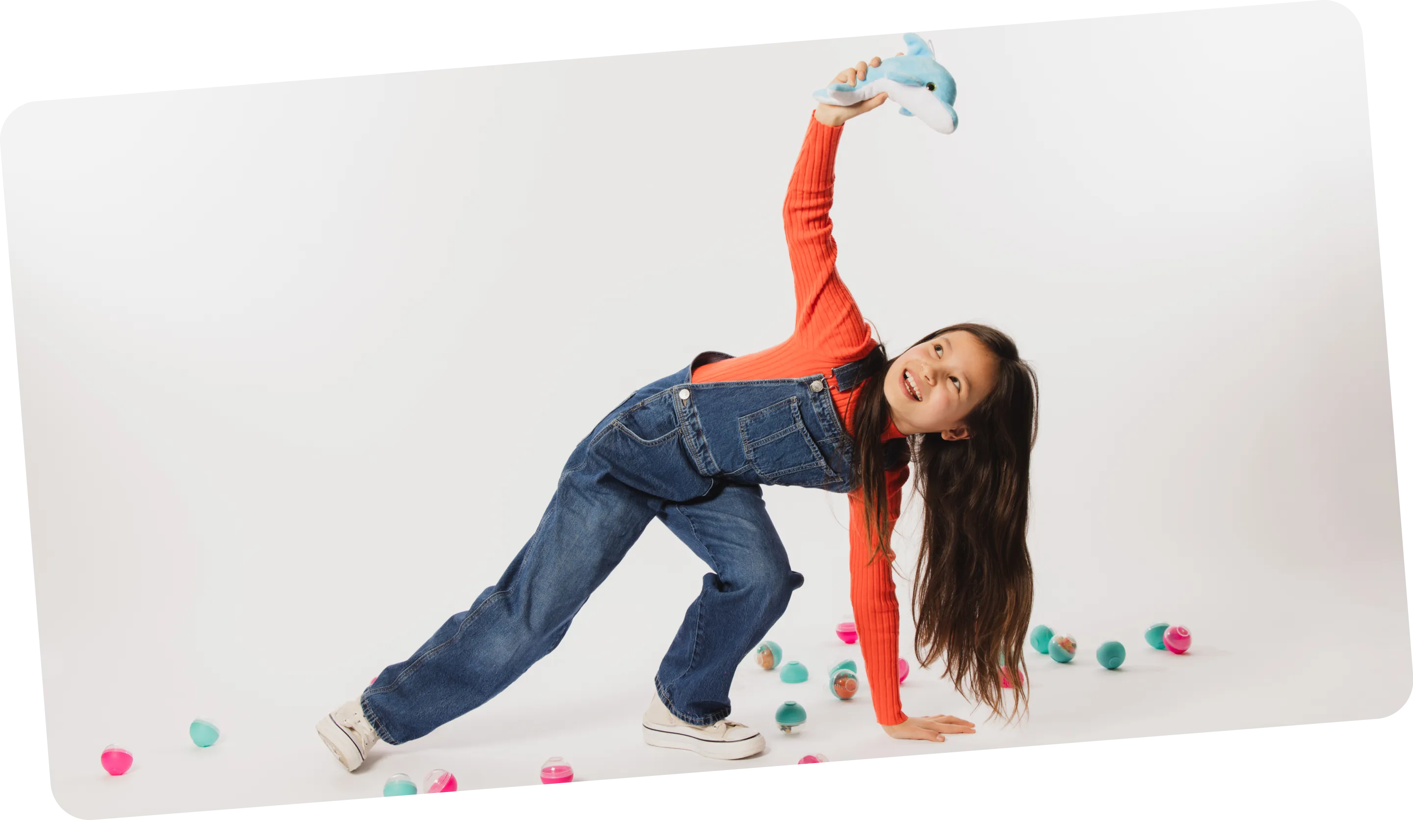Young girl in denim overalls and orange sweater playing with a blue stuffed dolphin, surrounded by colorful plastic capsules on a white background.