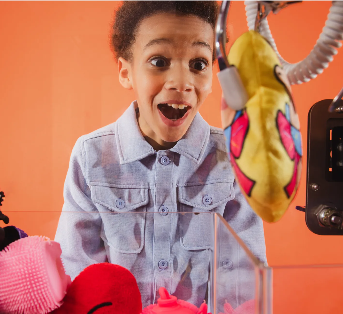 Excited boy wearing a light blue jacket watching a claw machine grabbing a yellow plush toy against an orange background.