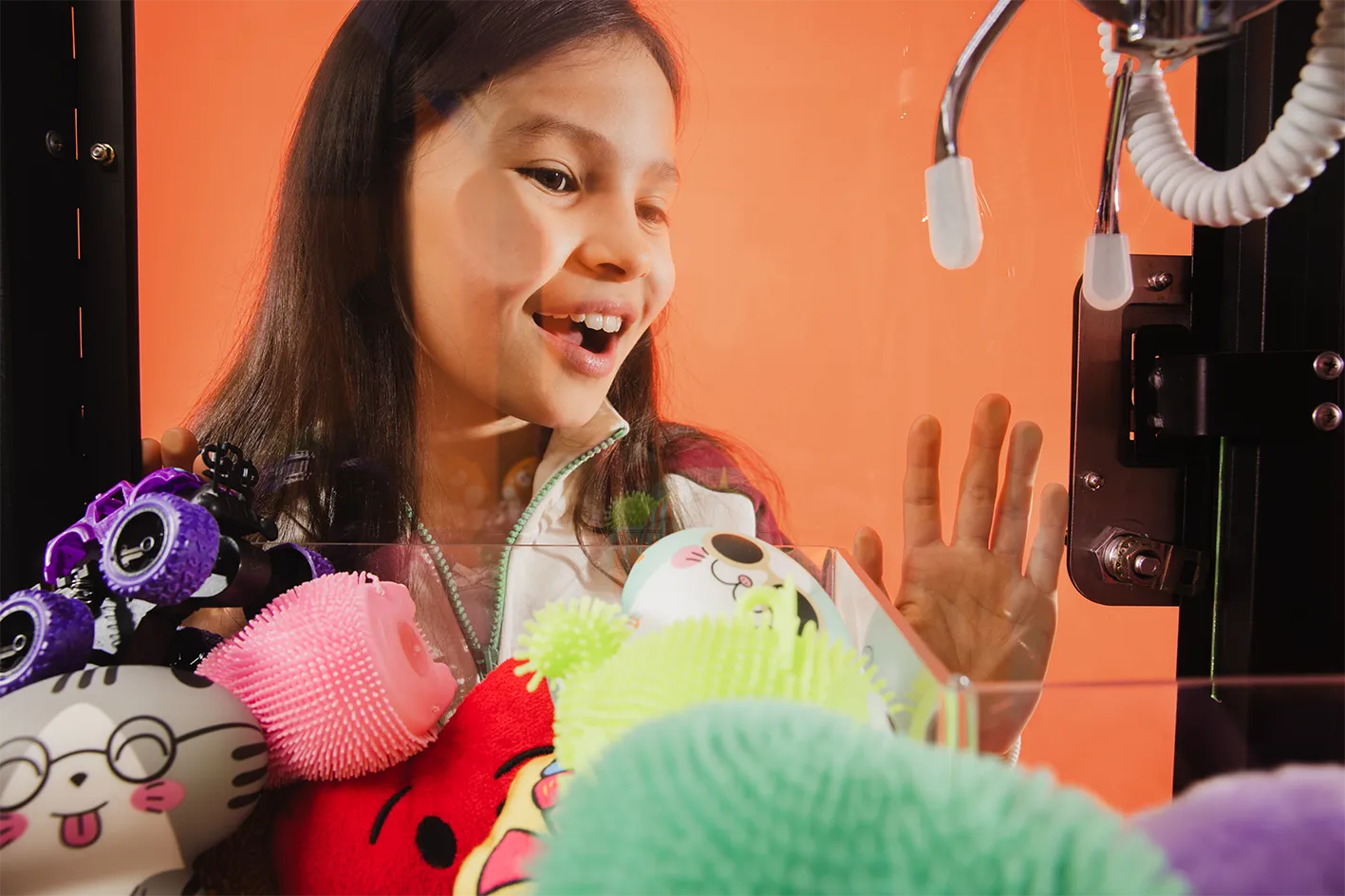 Smiling girl looking excitedly at plush toys inside a claw machine against an orange background.