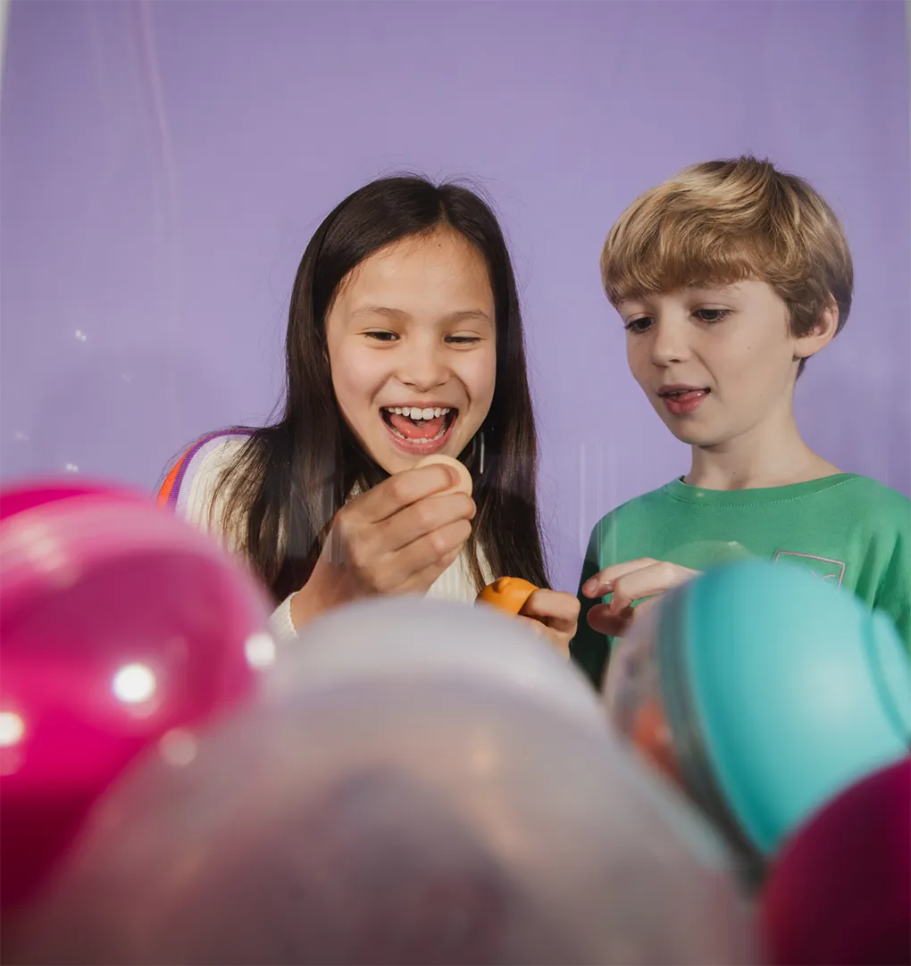 A smiling girl and a boy excitedly opening colorful plastic capsules against a purple background.