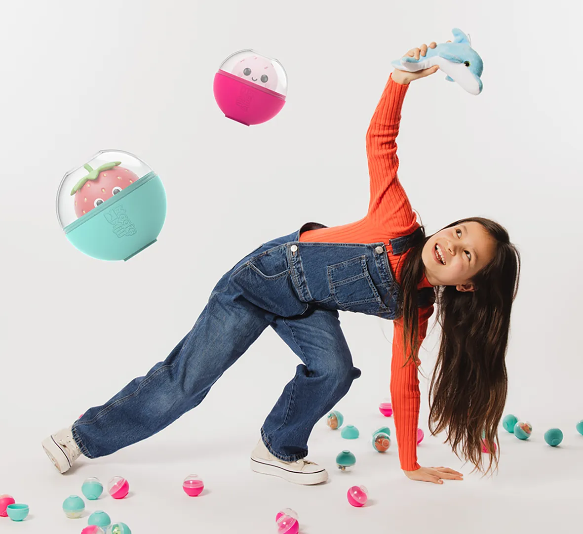 Smiling girl in denim overalls and orange sweater holding a blue dolphin toy, surrounded by colorful capsule toys with faces.
