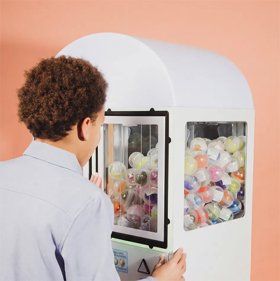 Person playing a white capsule toy vending machine filled with colorful plastic capsule balls against a peach background.