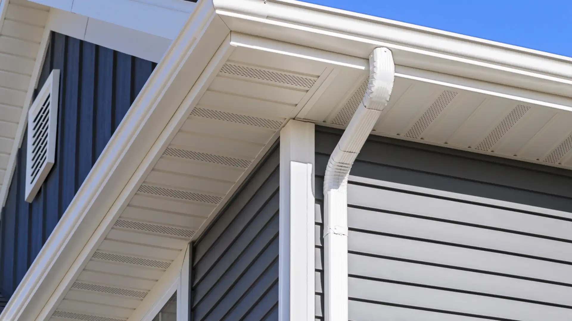Close-up of white gutter and soffit on gray vinyl siding under blue sky