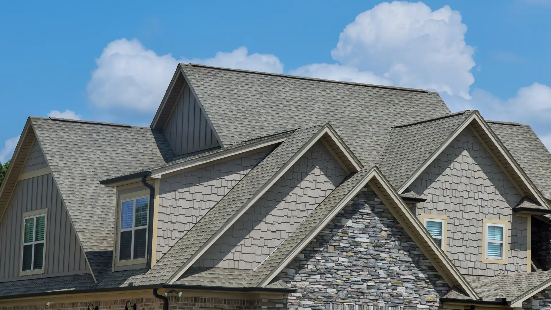 Residential home roof with gray asphalt shingles and stone exterior accents under blue sky