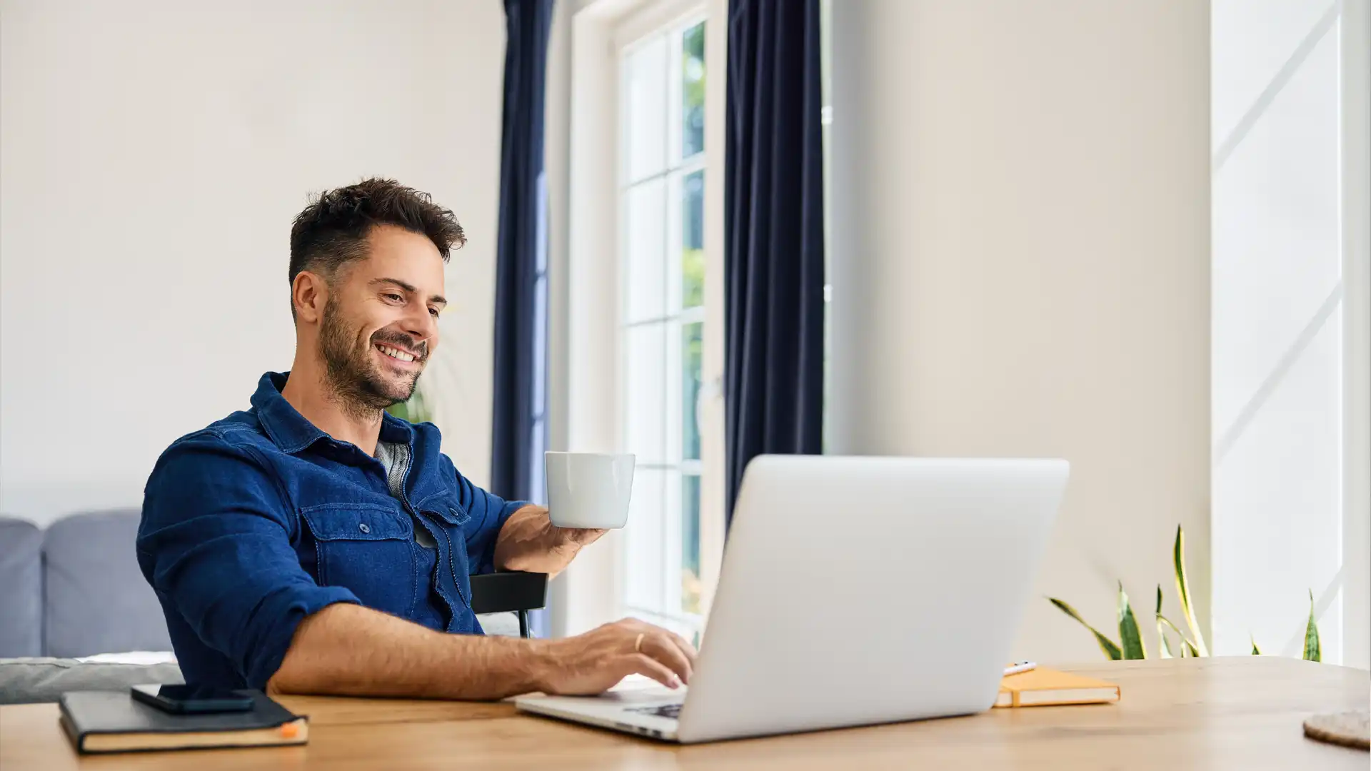 Smiling homeowner using a laptop at home while holding a coffee cup