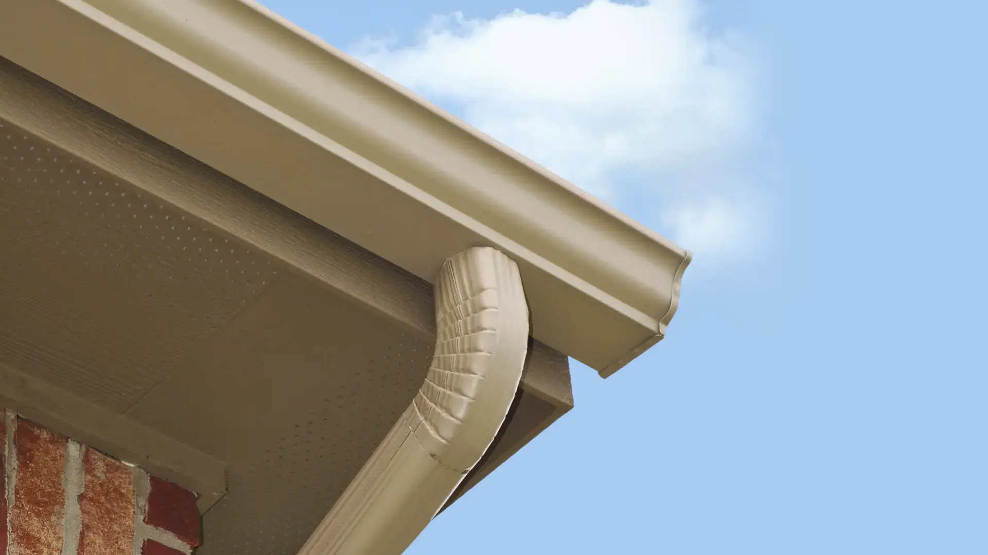 Close-up of beige gutter and downspout connection under roofline against blue sky.