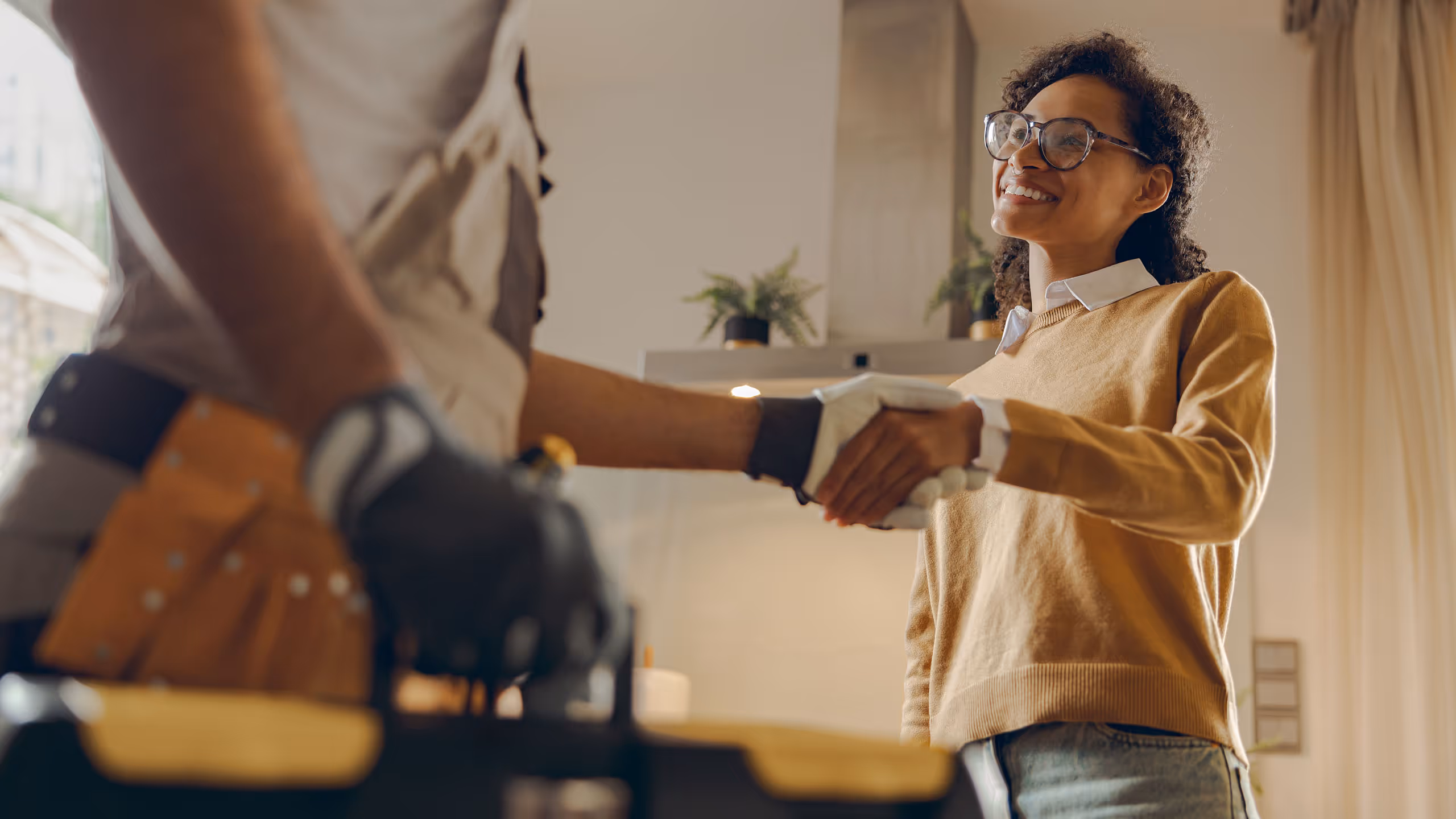 Smiling woman shaking hands with a worker wearing a tool belt and gloves indoors.