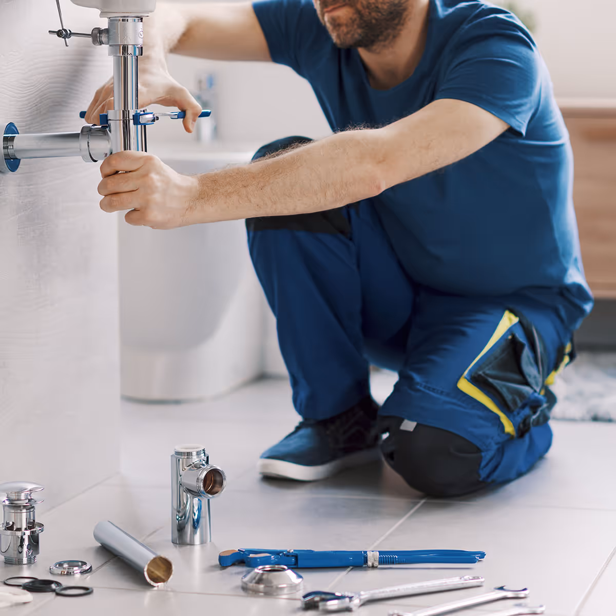 Plumber in blue uniform kneeling and tightening a pipe under a sink with plumbing tools laid out on tiled floor.