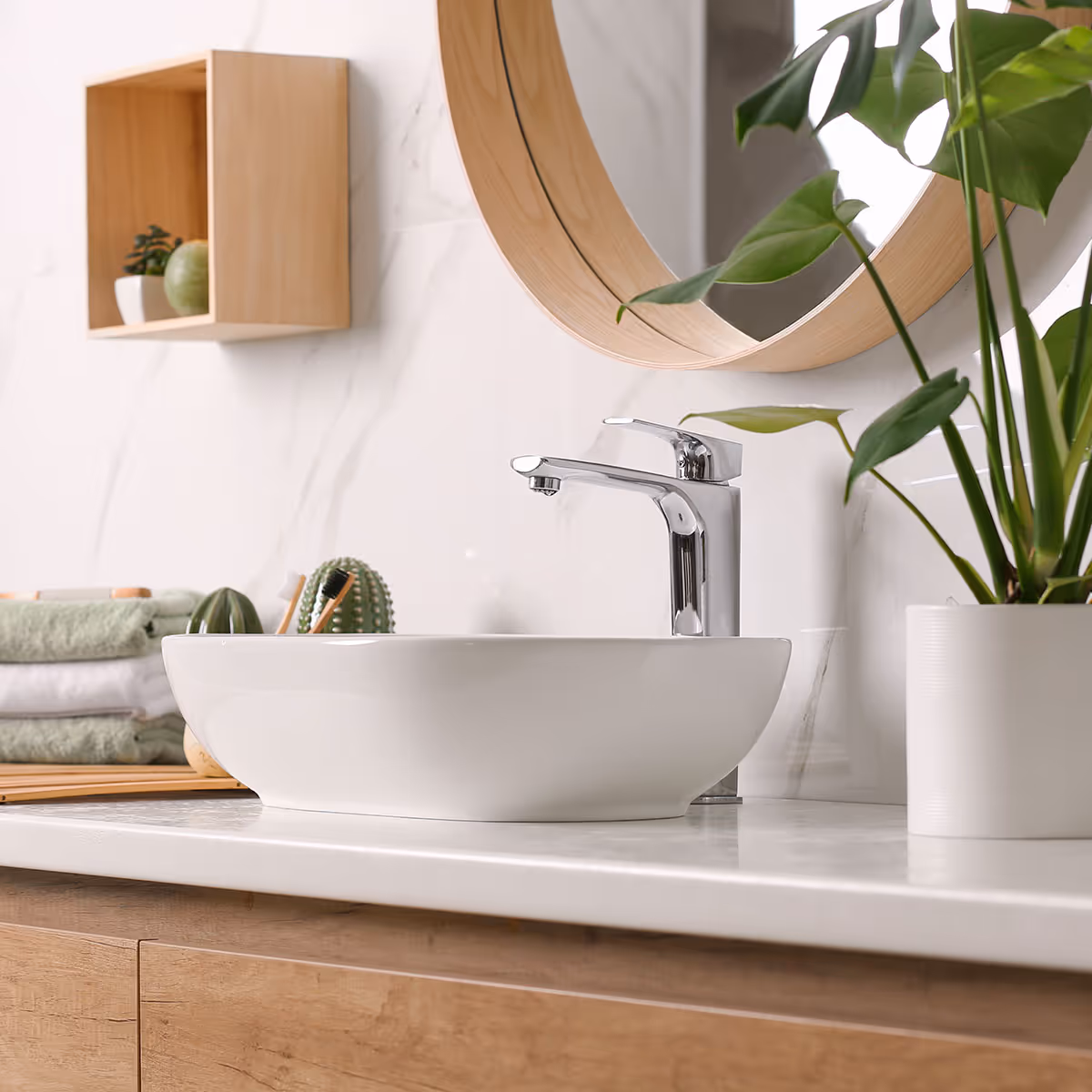 Modern bathroom sink with a chrome faucet, round wooden-framed mirror, potted green plant, and folded towels on a white countertop with wooden cabinetry.