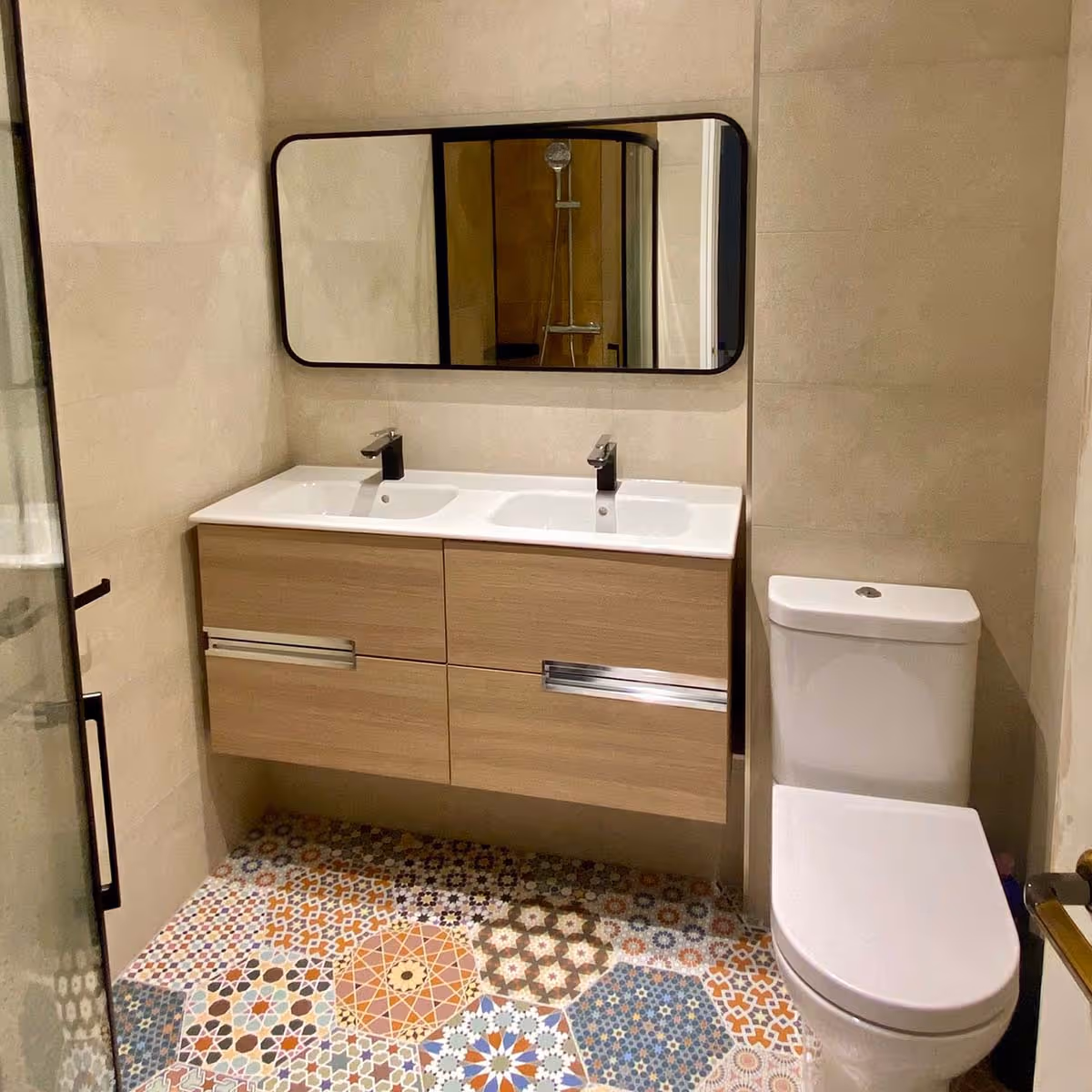 Modern bathroom with patterned tile floor, wall-mounted wooden vanity with double sinks and black faucets, rectangular mirror, and white toilet.