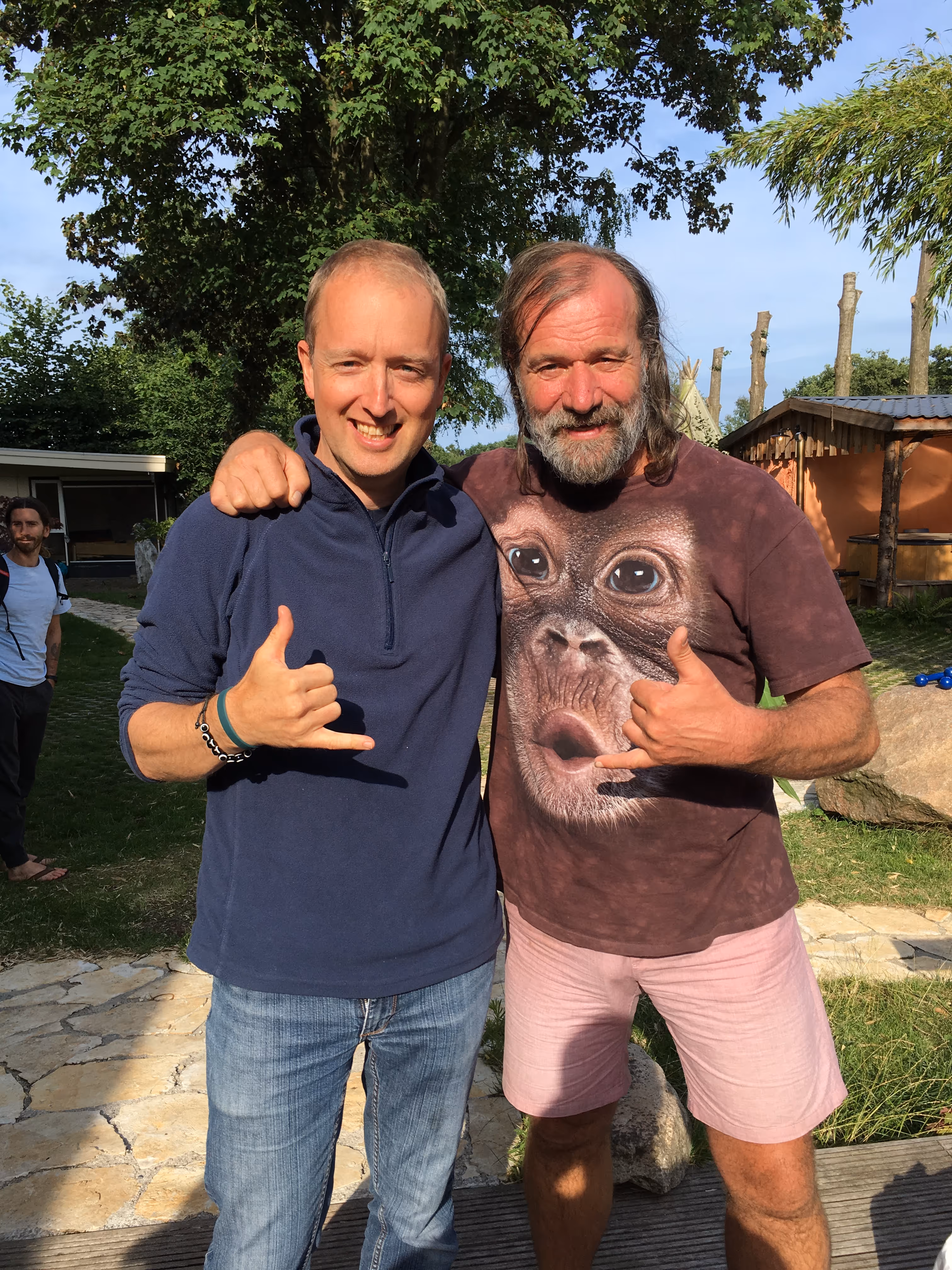 Deux hommes souriants posent dehors avec les bras autour de l’épaule, chacun faisant le geste de la main en shaka.