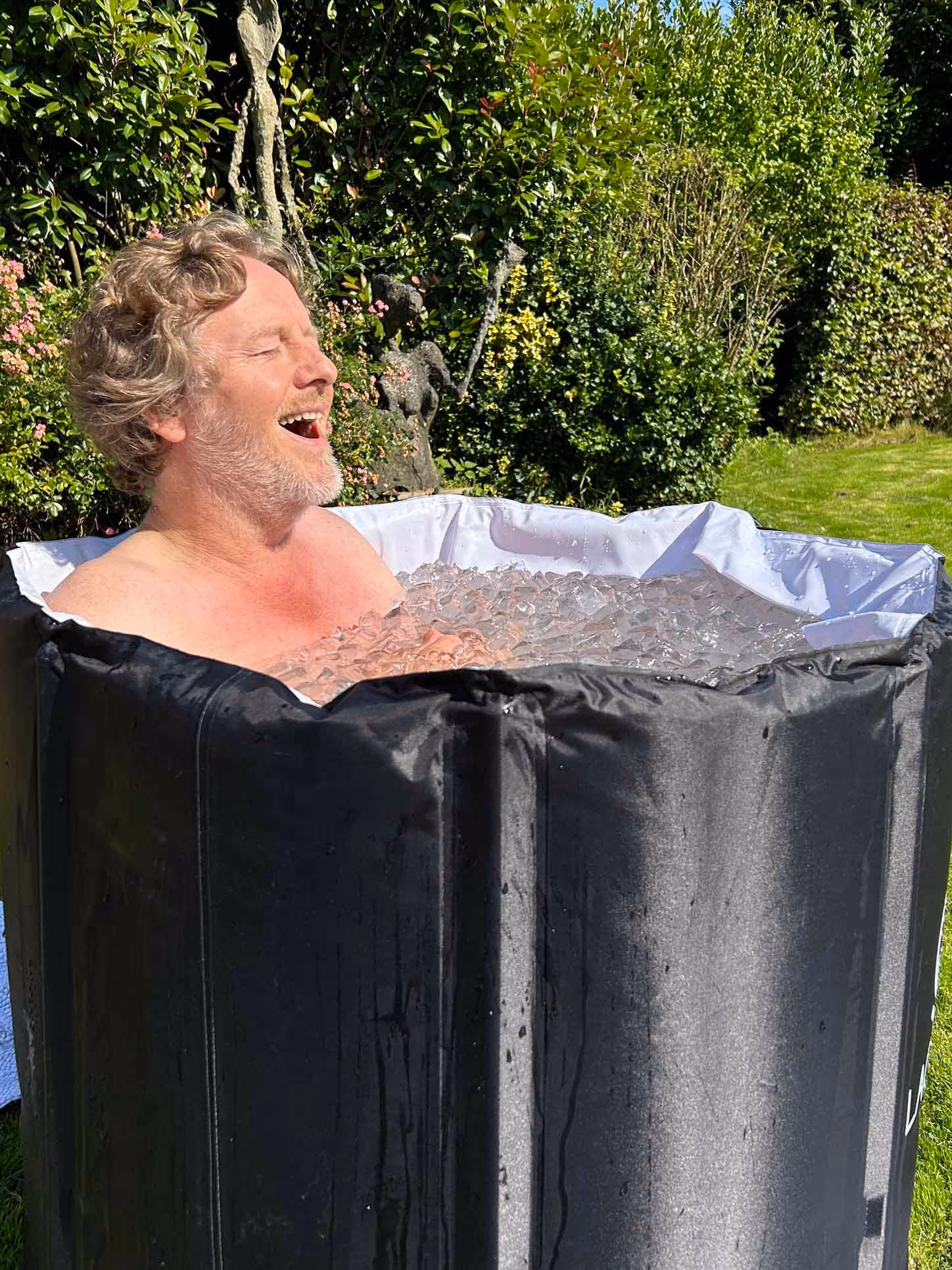 Un homme aux cheveux bouclés est assis dans un bain de glace extérieur, semblant ressentir le froid avec les yeux fermés et la bouche ouverte.