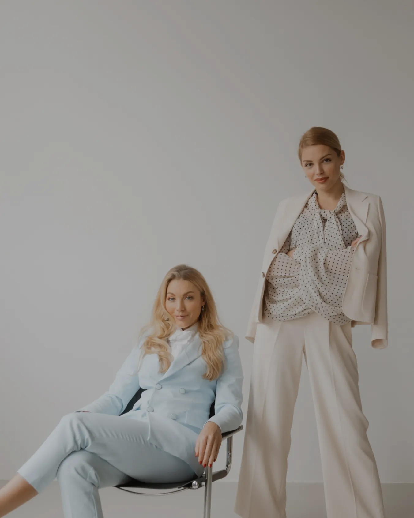 Zwei elegant gekleidete Frauen, eine sitzt in einem Stuhl im hellblauen Anzug, die andere steht im cremefarbenen Anzug mit gepunkteter Bluse vor hellgrauem Hintergrund.
