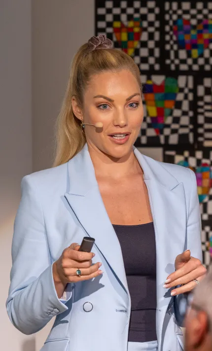 Frau mit blonder Pferdeschwanz-Frisur und hellem Anzug hält eine Fernbedienung und spricht mit Headset-Mikrofon vor buntem Schachbrettmuster.