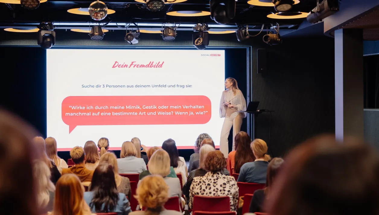 Frau hält Präsentation vor Publikum, zeigt Folie mit deutscher Frage zu Mimik, Gestik und Verhalten.