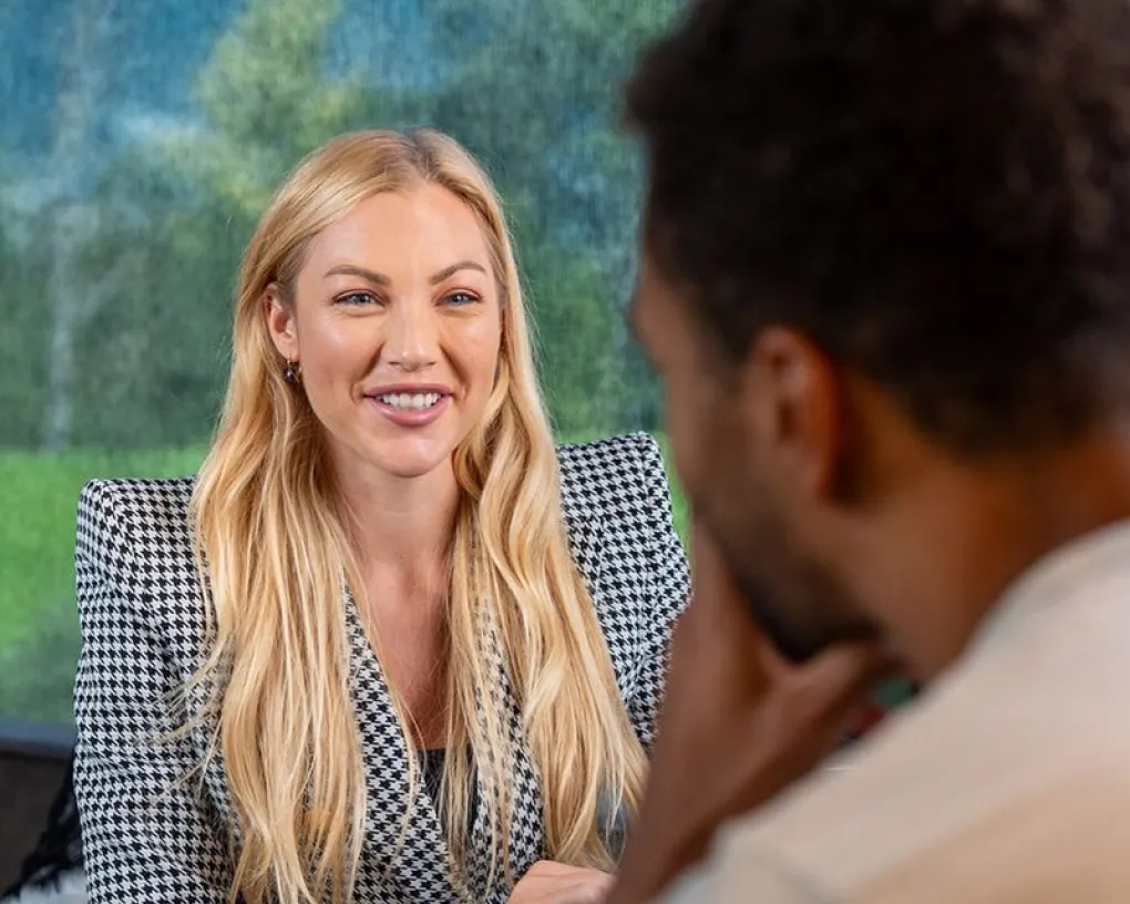 Eine lächelnde blonde Frau trägt einen schwarz-weißen Hahnentritt-Blazer und führt ein Gespräch mit einem Mann im Vordergrund.