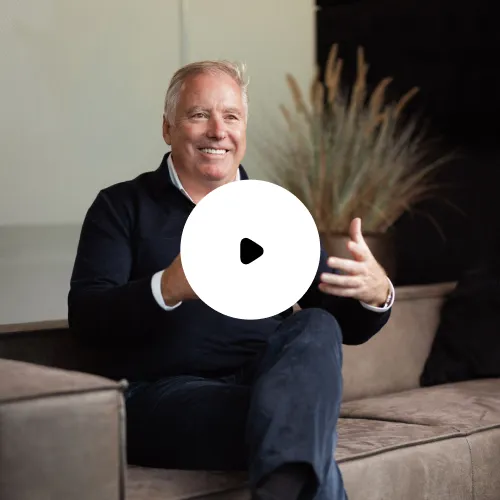 Älterer Mann mit grauem Haar, der auf einem Sofa sitzt und lächelt, während er gestikuliert.