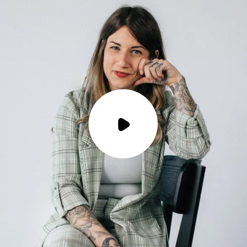 Frau in kariertem Anzug mit Tätowierungen, die auf einem Stuhl sitzt und in die Kamera lächelt.