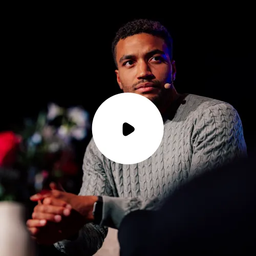 Mann mit Mikrofon headset trägt grauen Strickpullover und hört aufmerksam zu, während er auf einer Bühne spricht.