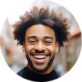 Smiling man with curly hair and a beard, wearing a black shirt, outdoors with blurred background.