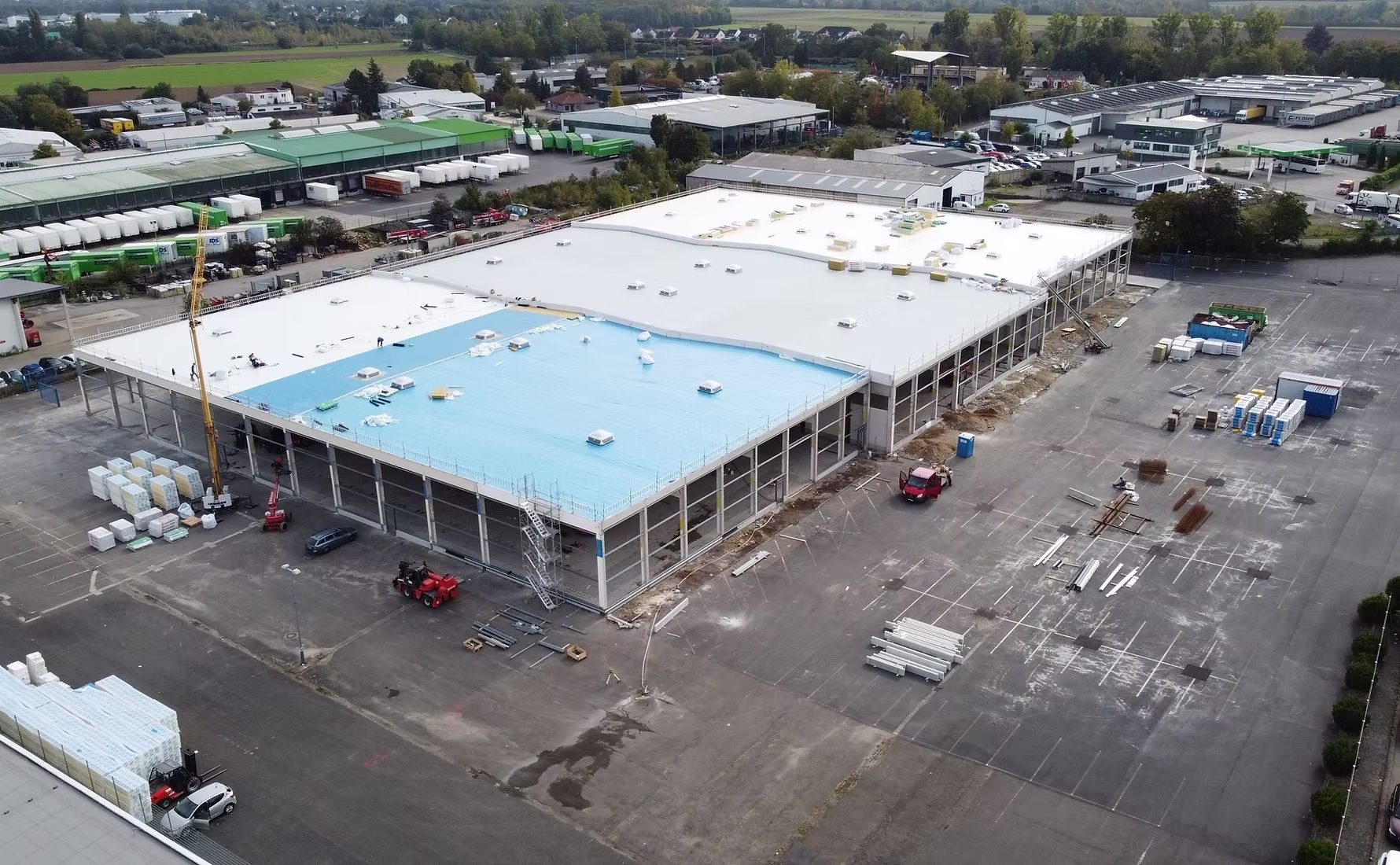 Luftaufnahme einer großen Lagerhalle im Bau mit blauem und weißem Dach und Baustoffen davor.