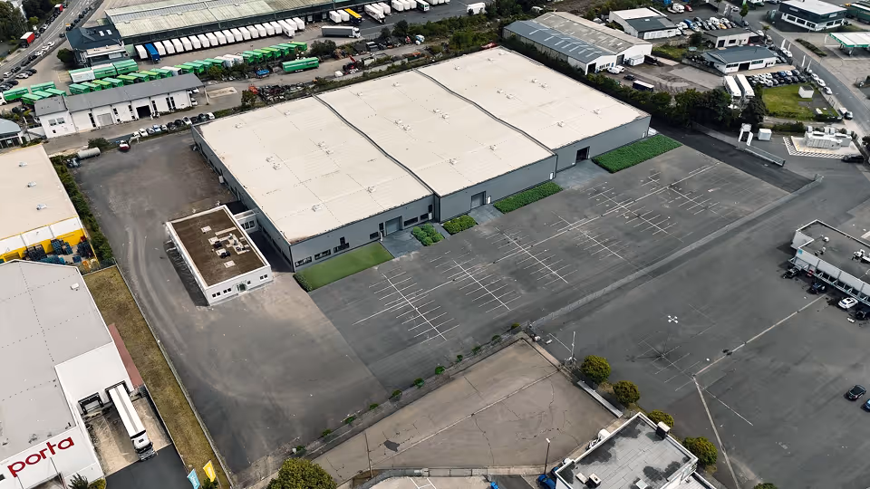 Luftaufnahme eines großen Industriegebäudes mit leerem Parkplatz und benachbarten Lagerhallen.