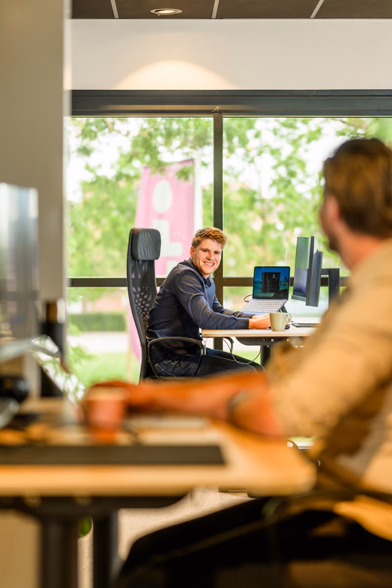Een glimlachende man werkt op een laptop aan een bureau naast een raam in een kantoor.