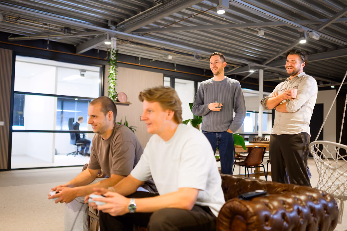 Vier mannen spelen en kijken lachend naar een videogame in een moderne kantooromgeving.