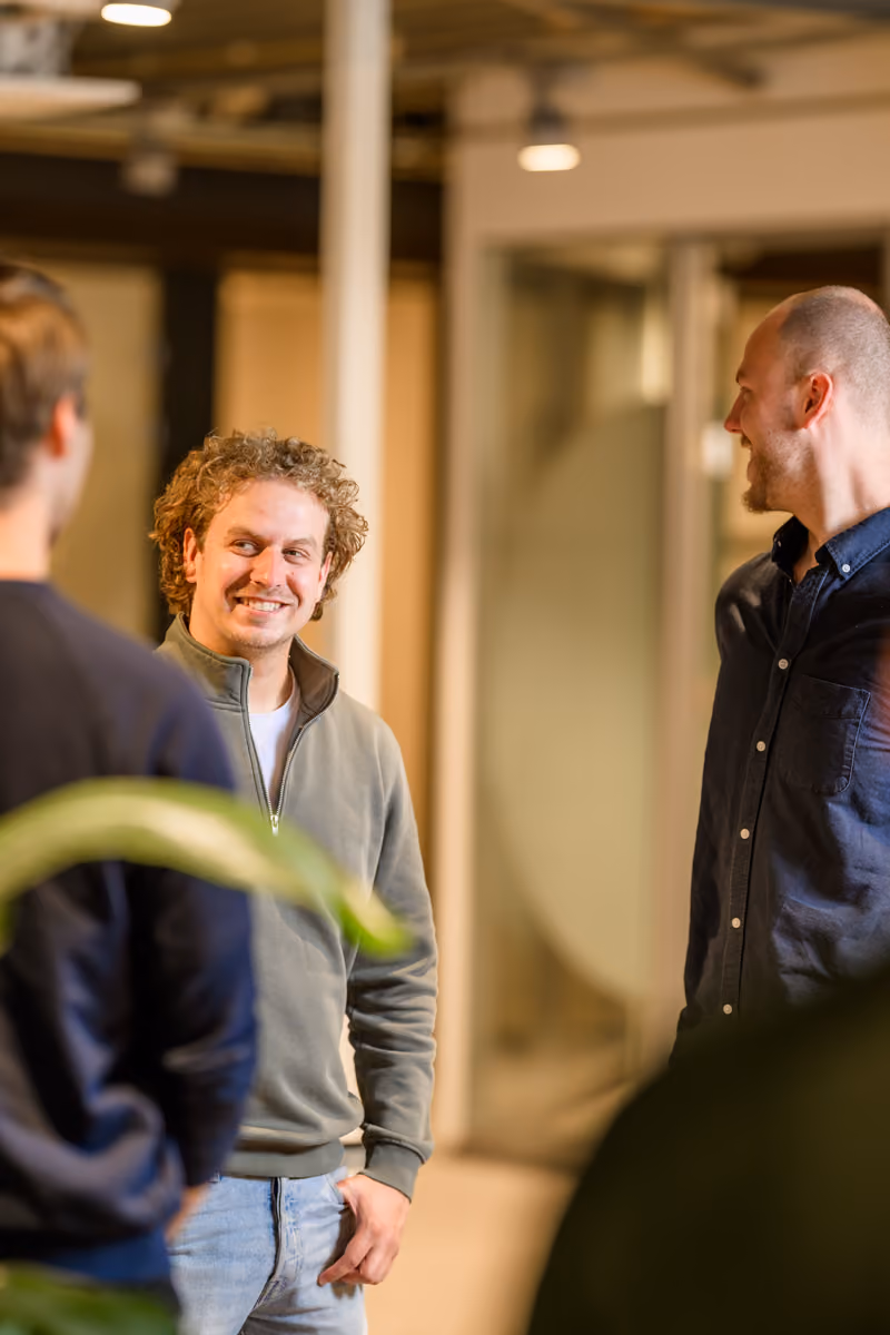 Drie mannen staan in een kantoorruimte en praten met elkaar, twee kijken lachend naar een derde.