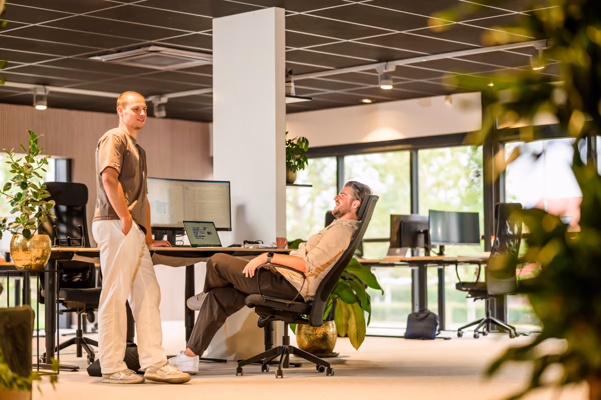Twee mannen ontspannen en praten in een lichte moderne kantooromgeving met planten en computers.