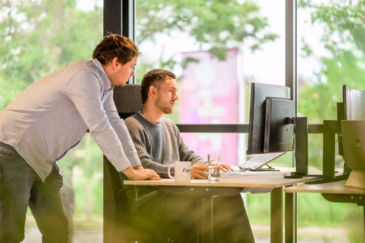 Twee mannen werken samen bij een bureau met een computerscherm in een kantoor met grote ramen.