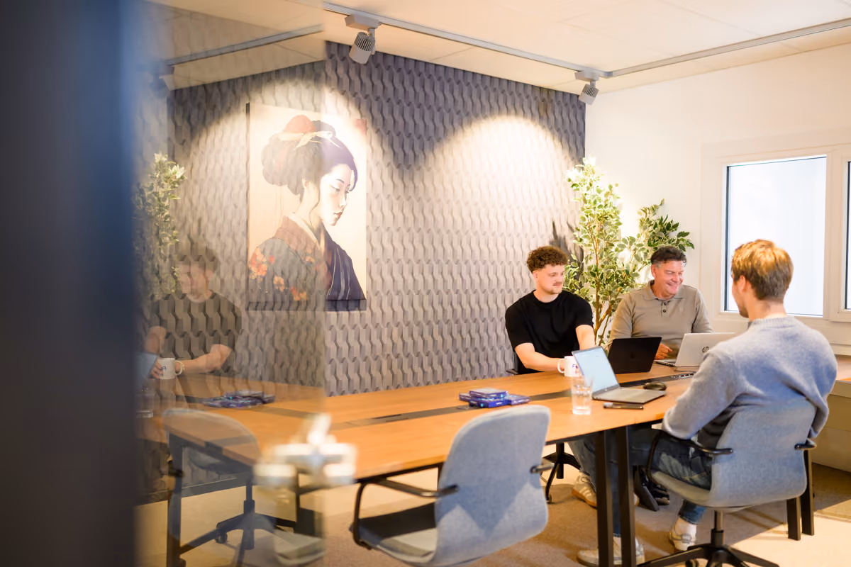 Drie mannen zitten samen aan een vergadertafel met laptops en koffiekopjes in een lichte kamer.