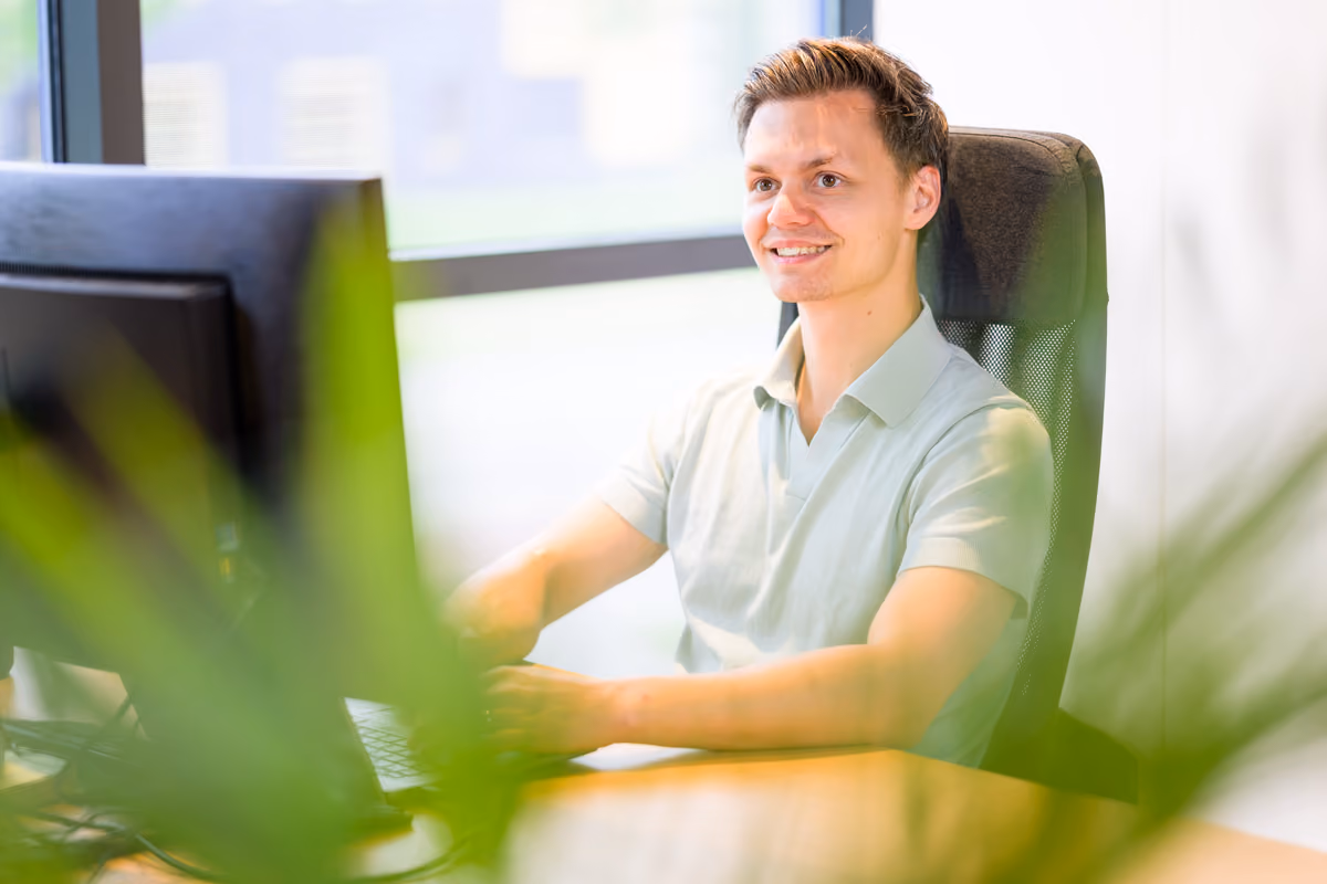 Jonge man die glimlacht terwijl hij aan een bureau aan een computer werkt in een helder kantoor.