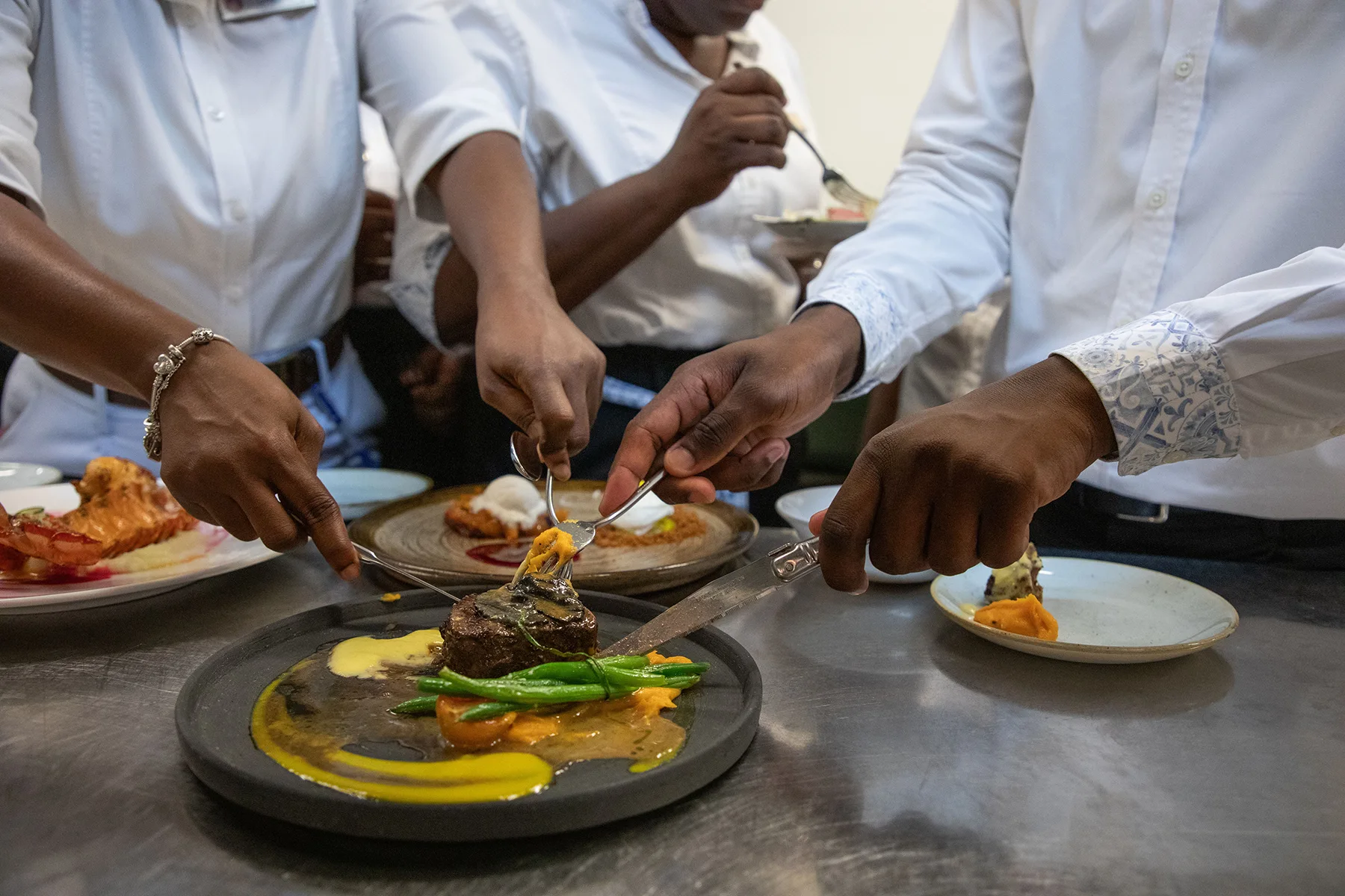 Three people in white shirts serving gourmet food from a black plate with steak, green beans, and sauce onto smaller plates.