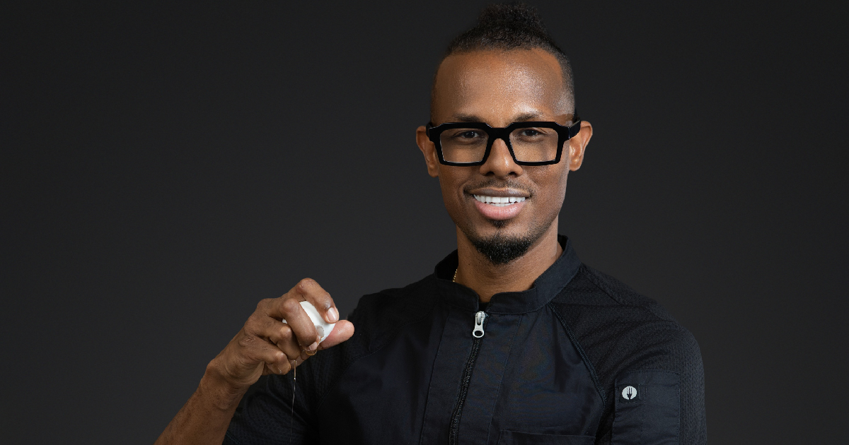 Smiling man wearing black chef jacket and glasses cracking an egg against a dark background.
