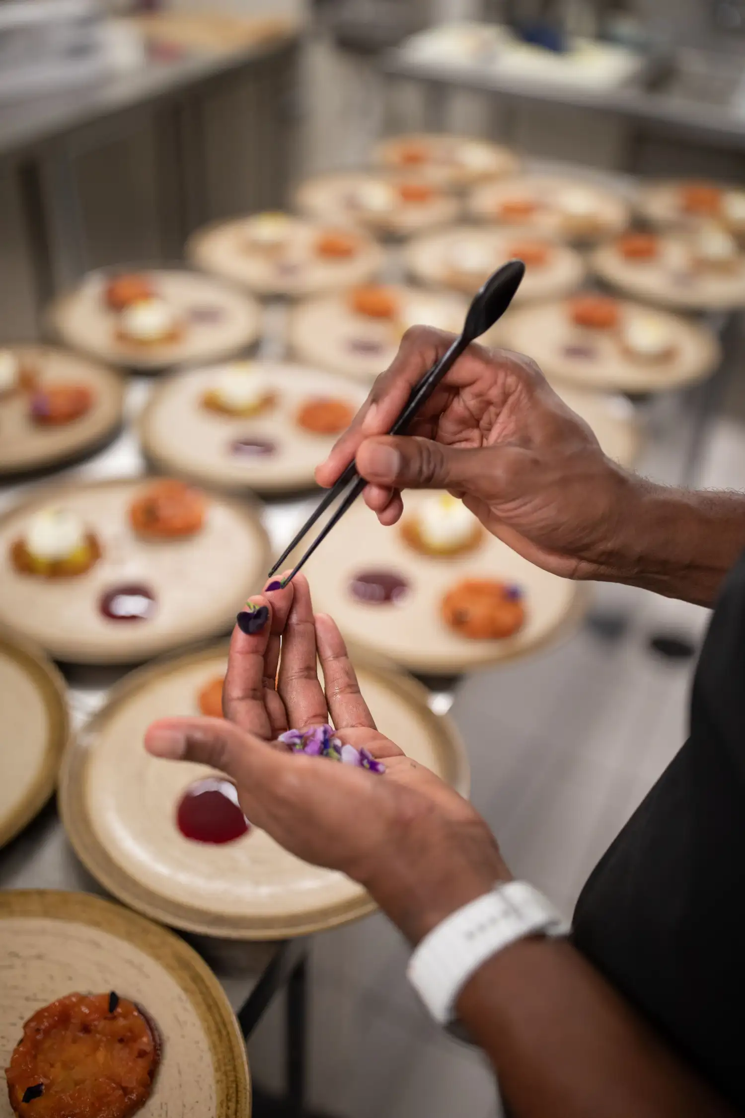 Chef using tweezers to carefully place edible purple flowers on a dish in a professional kitchen.