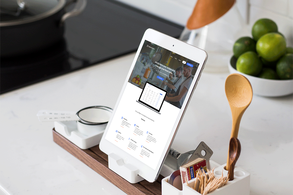 Tablet on wooden stand displaying a food management website, surrounded by kitchen utensils on a white countertop with a bowl of limes in the background.