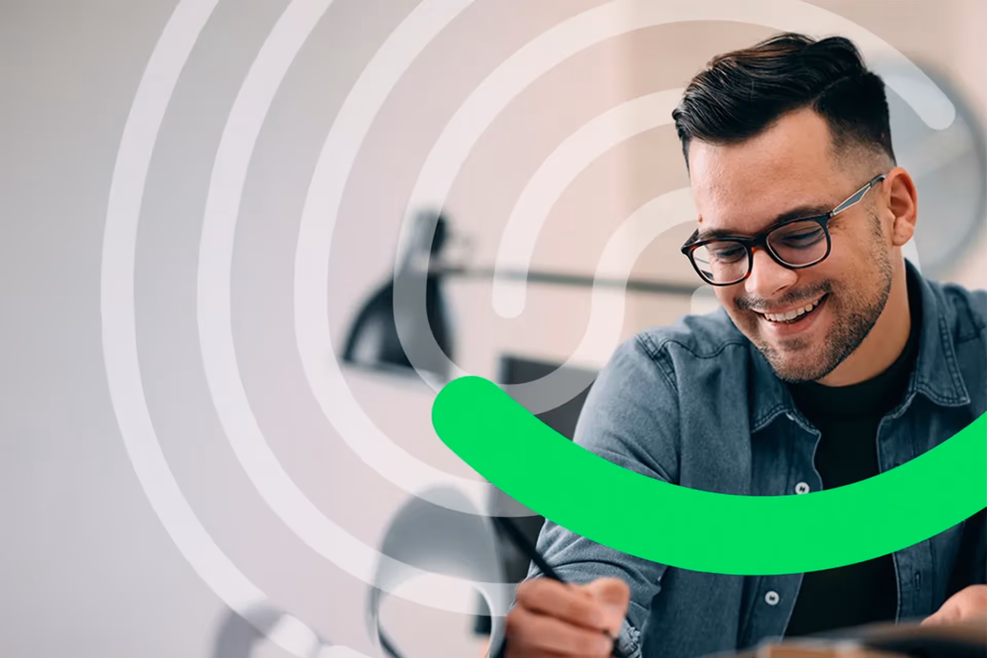 Smiling man with glasses writing or drawing at a desk in a modern office setting.